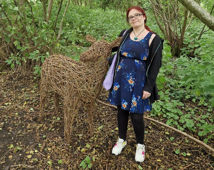 A woman standing next to a chest-height model deer made out of sticks, in woodland