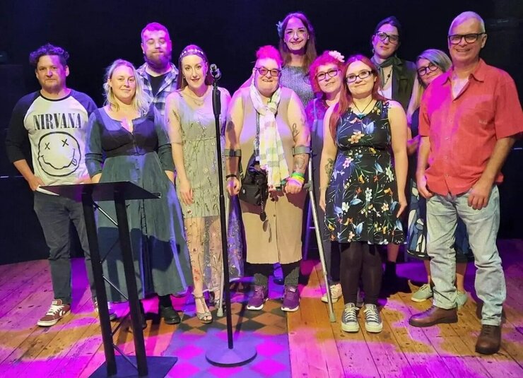 Eleven people standing on a wooden floor with a microphone, under stage lighting